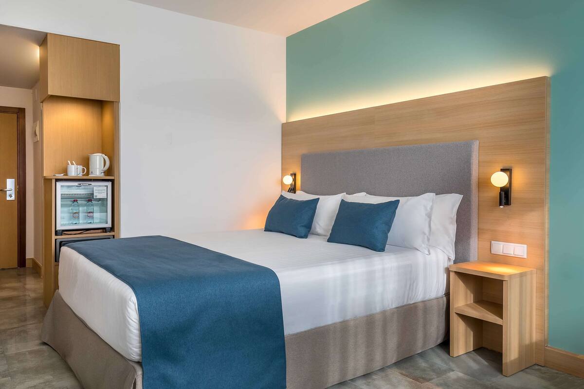 A hotel room with a large bed, bedside tables, a minibar, and a wall-mounted light fixture.