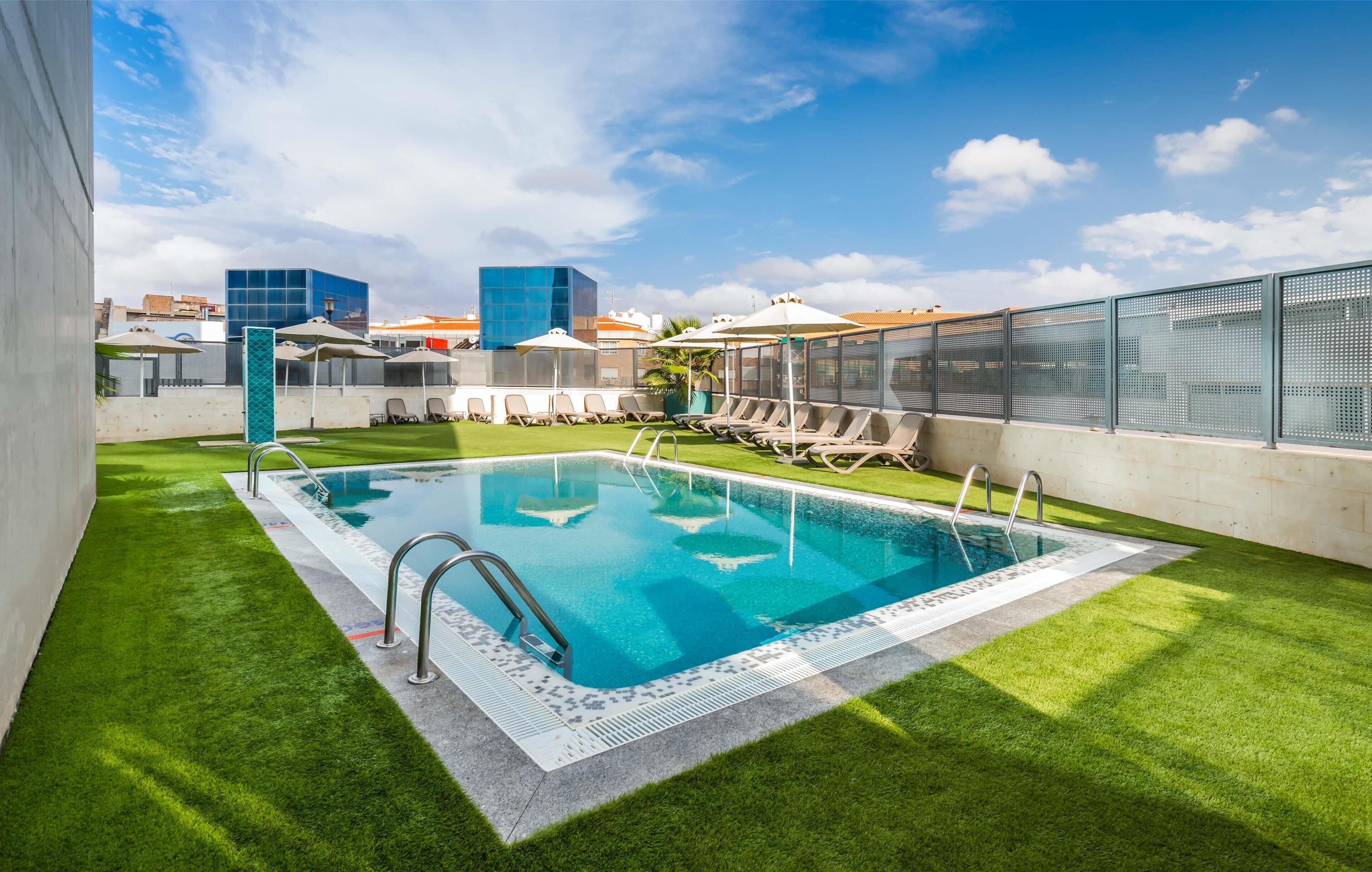 Una piscina con agua azul cristalina, rodeada de césped artificial y una valla con diseño de malla.