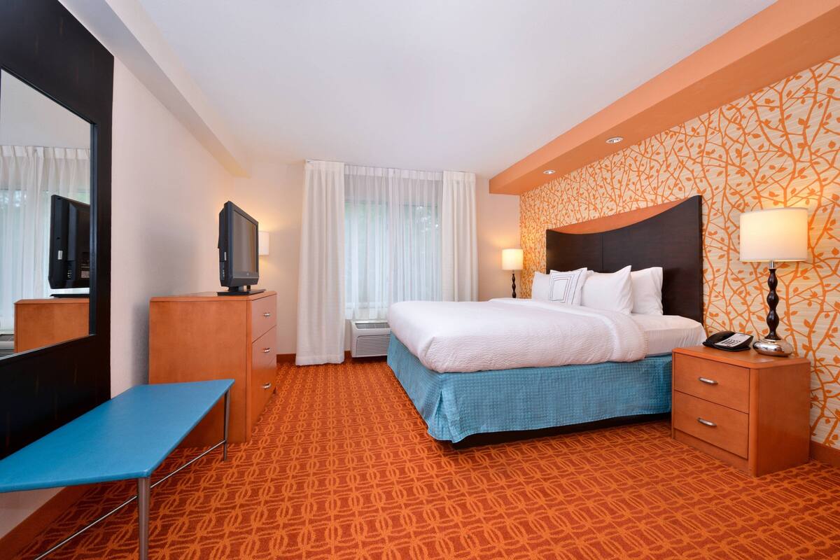 A hotel room with a bed, bedside table, lamp, and a television on a cabinet.