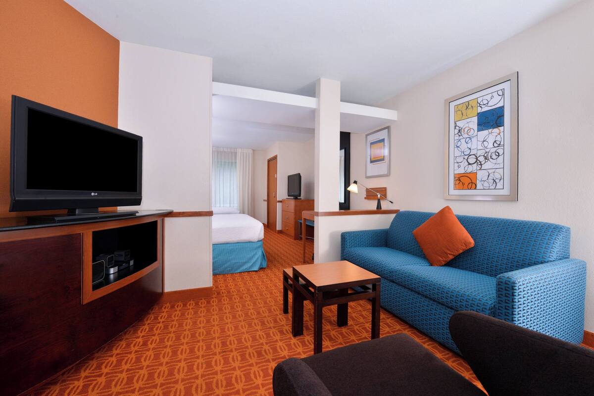 A hotel room with a TV, a blue sofa, a small table, a bed, and a desk with a lamp.