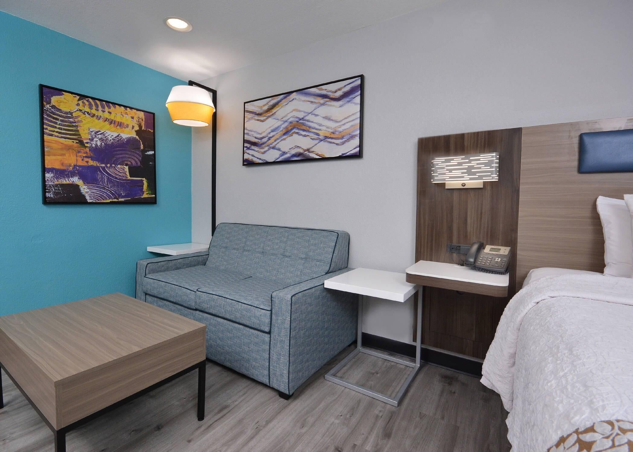 Une chambre d’hôtel moderne avec un canapé gris, une table en bois, un bureau avec un téléphone et des œuvres d’art accrochées aux murs.