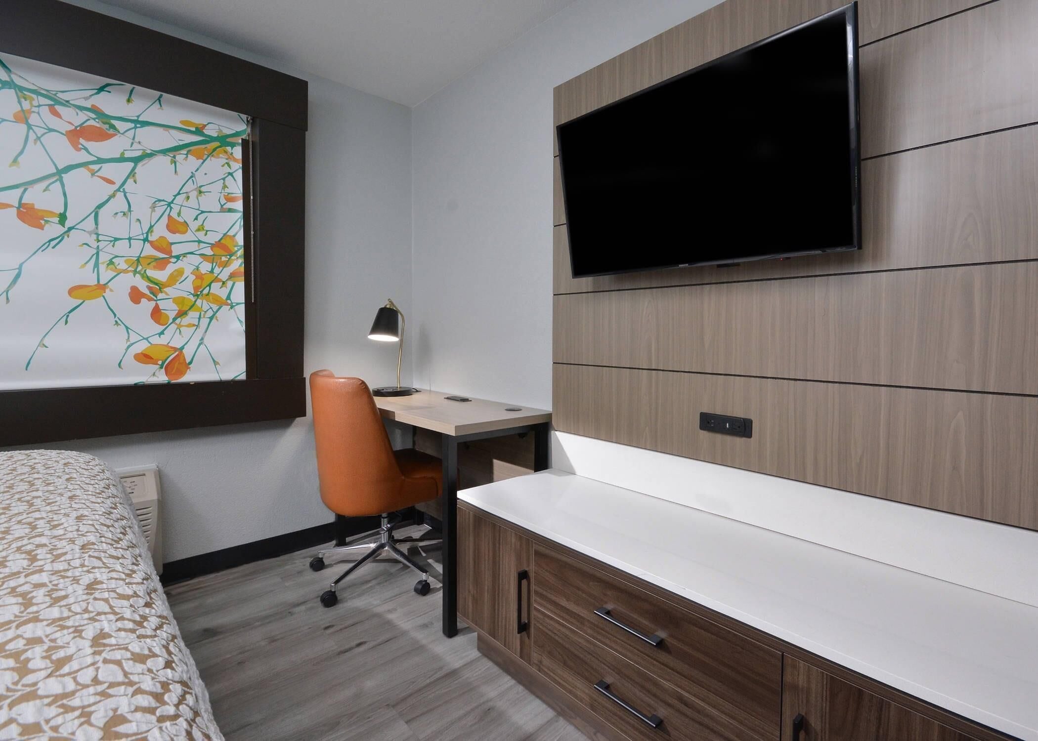 Habitación de hotel con cama, escritorio, sillón naranja, televisor montado en la pared y ventana grande con vista floral.