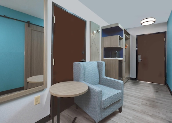 A hotel room with a blue armchair, a small round table, a wardrobe, and a kitchenette area.