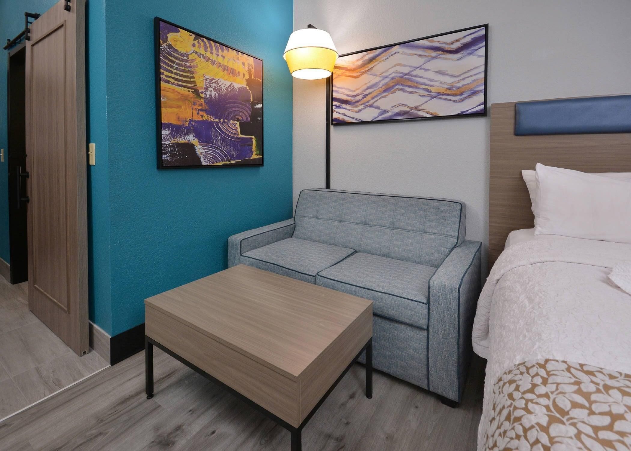 Ein Hotelzimmer mit grauem Sofa, einem Holztisch, einem Bett mit weißer Bettwäsche und zwei abstrakten Bildern an den Wänden.