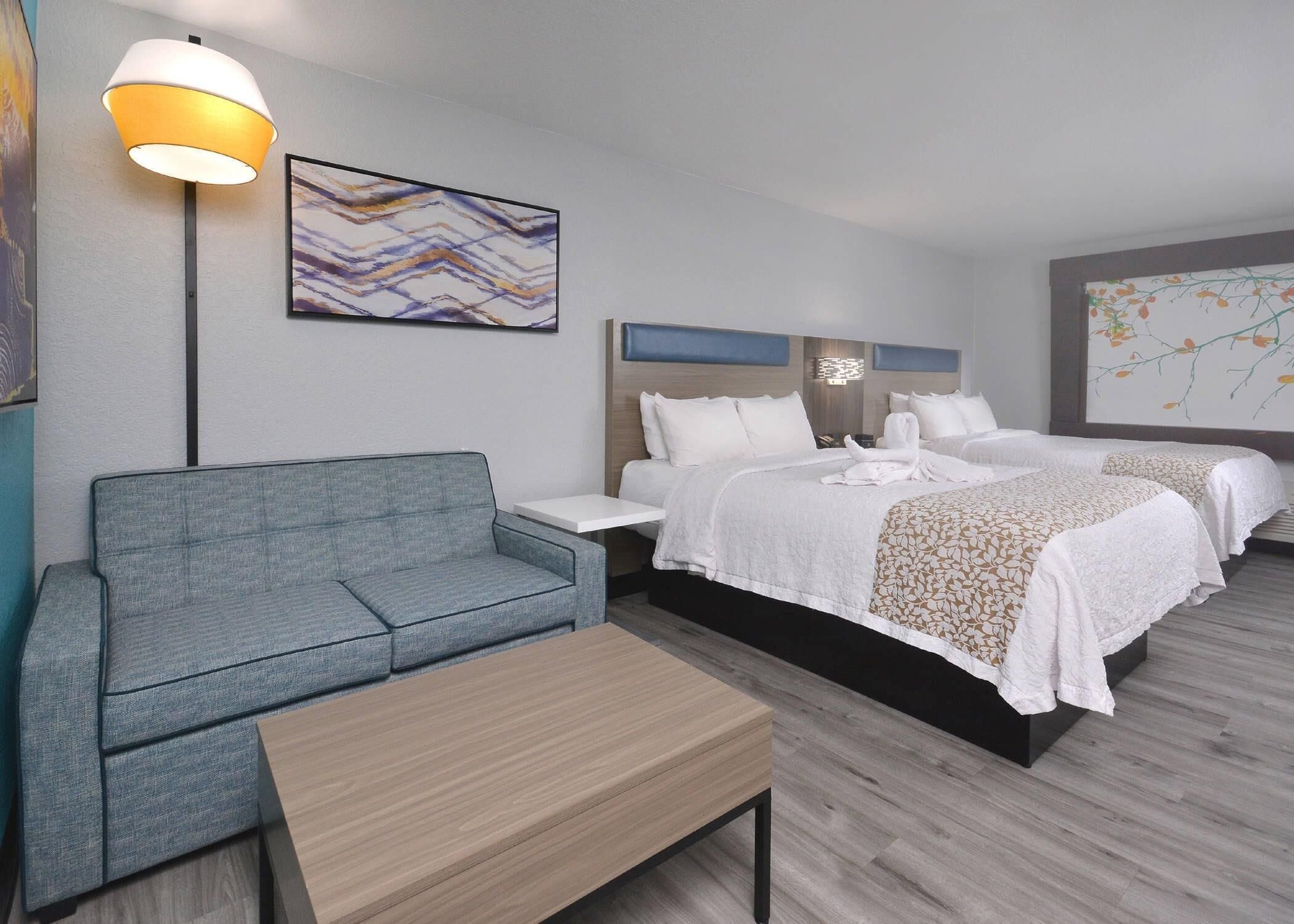 A hotel room with a grey sofa, a wooden coffee table, two beds with white linens, a wall-mounted lamp, and abstract art.