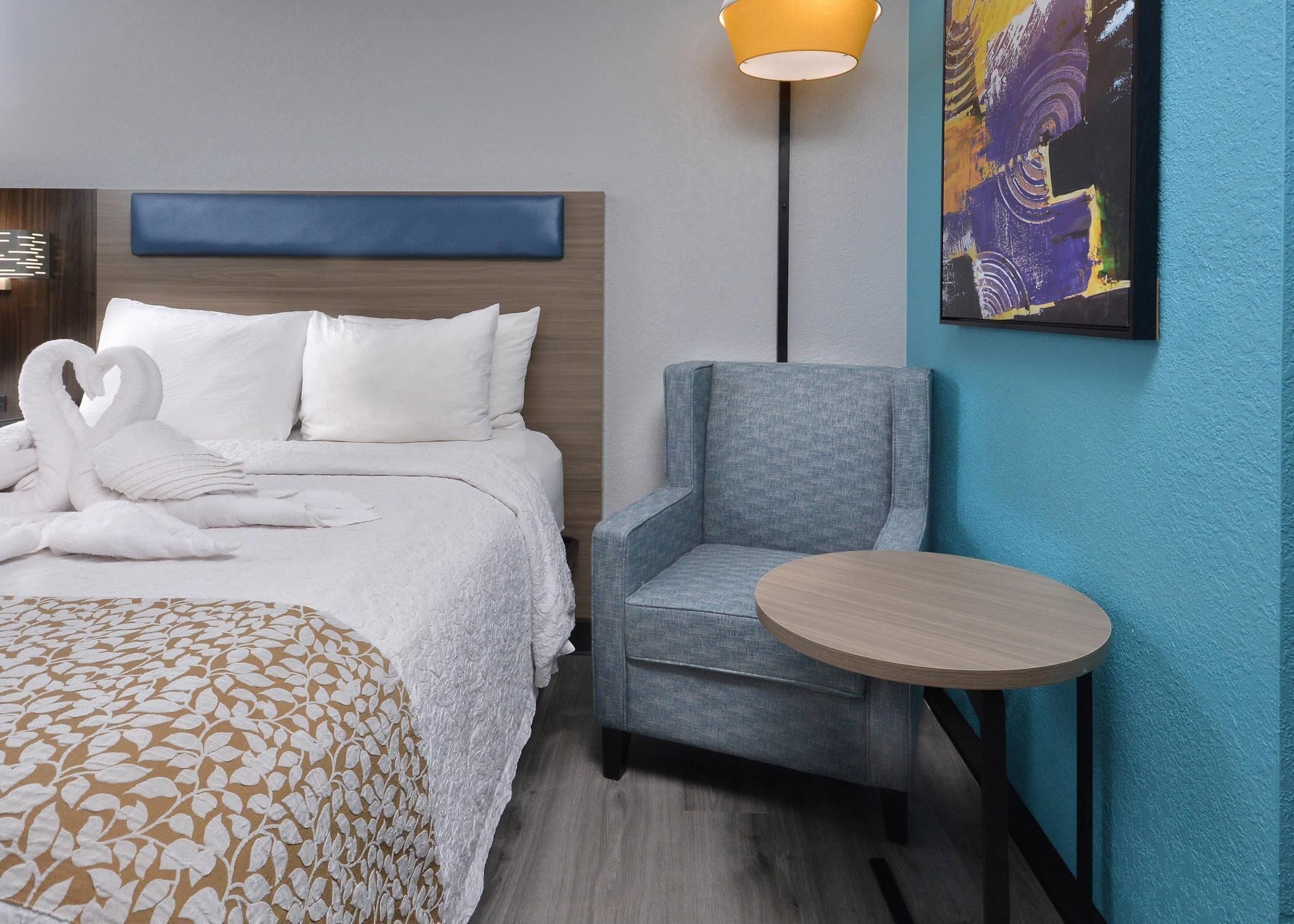 A hotel room with a neatly made bed, a blue upholstered armchair, a small round table, and a painting on the wall.