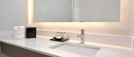 Une salle de bain avec un comptoir blanc, un lavabo, un miroir, un porte-serviettes et un rideau de douche.
