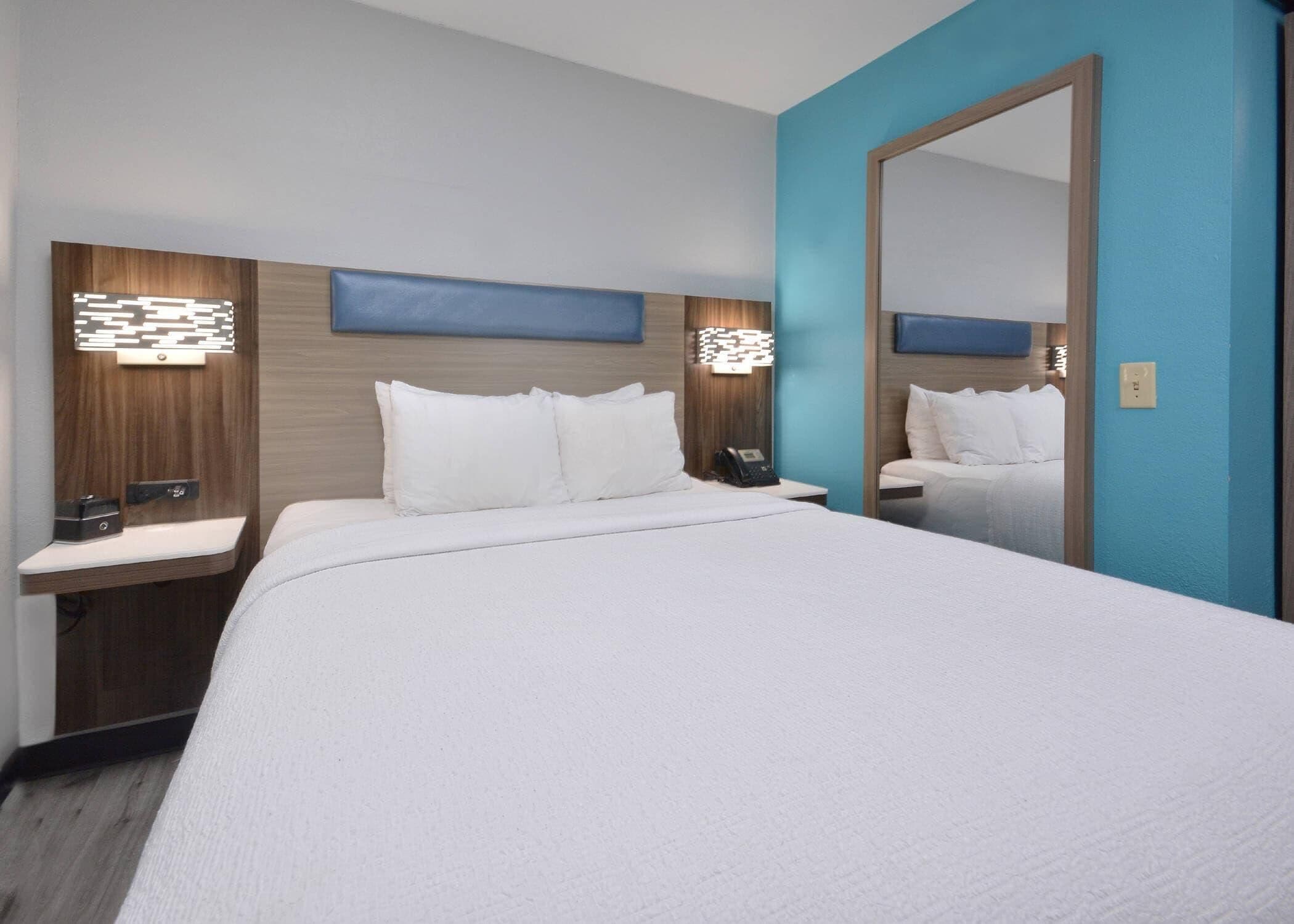 Una habitación de hotel con una cama grande, una mesita de noche, un espejo y una lámpara fijada a la pared.