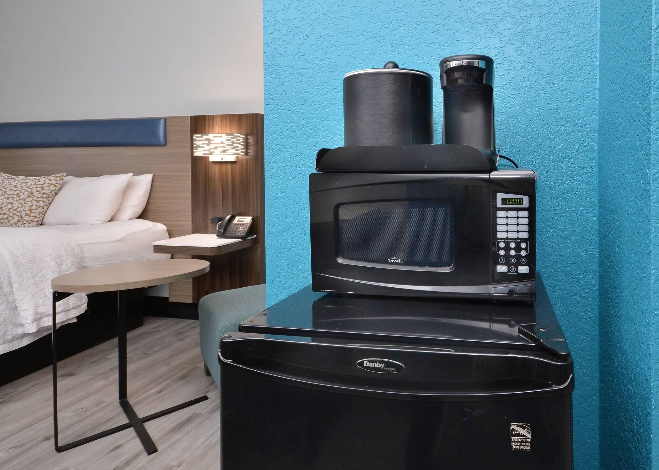A compact, black electric stove with a digital display and a coffee maker on top, placed in a modern hotel room with a blue accent wall, a wooden headboard, and a small round table with a telephone.