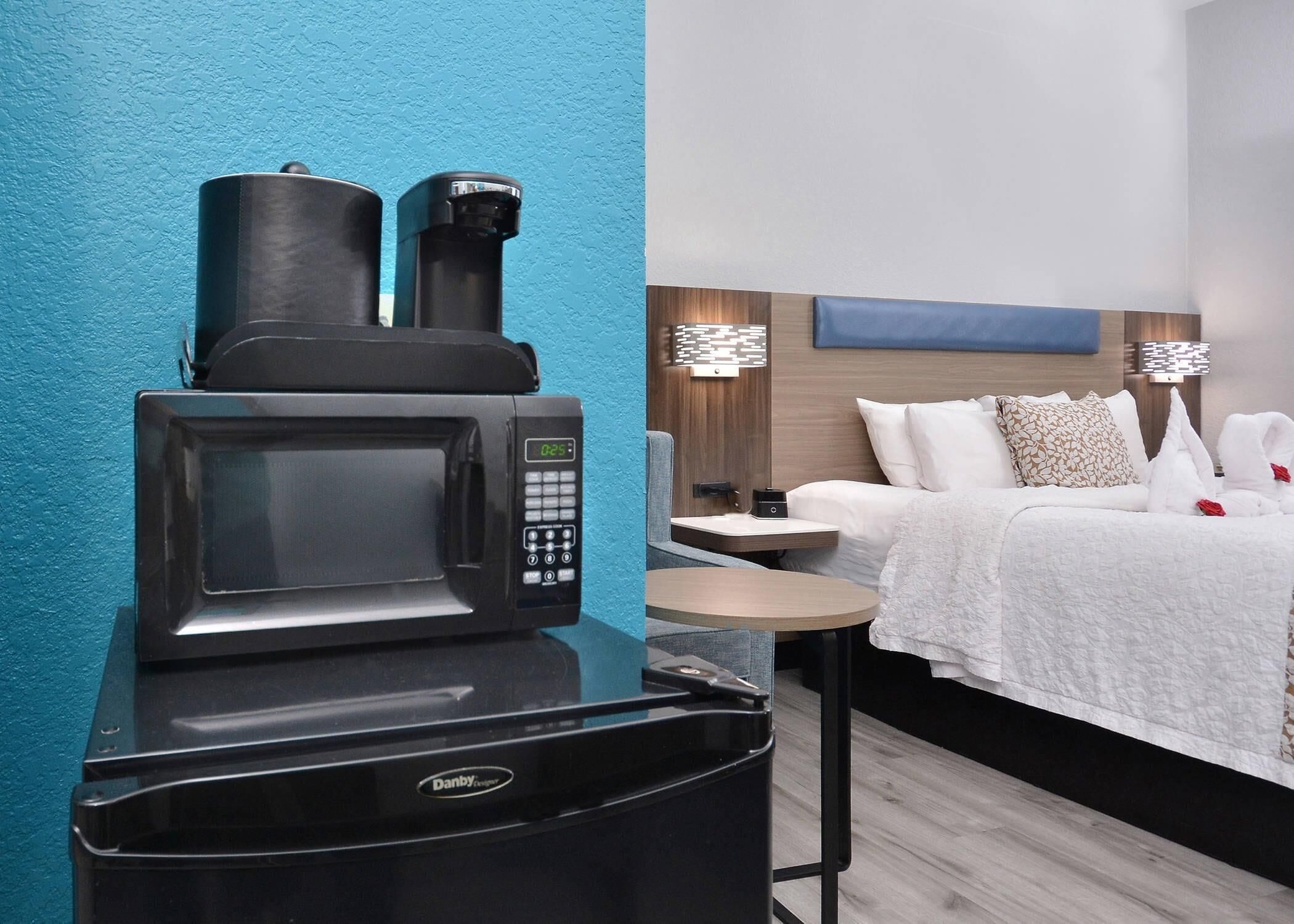 A compact room with a microwave and coffee maker on a counter, a small table, a bed with white bedding, and a blue accent wall.