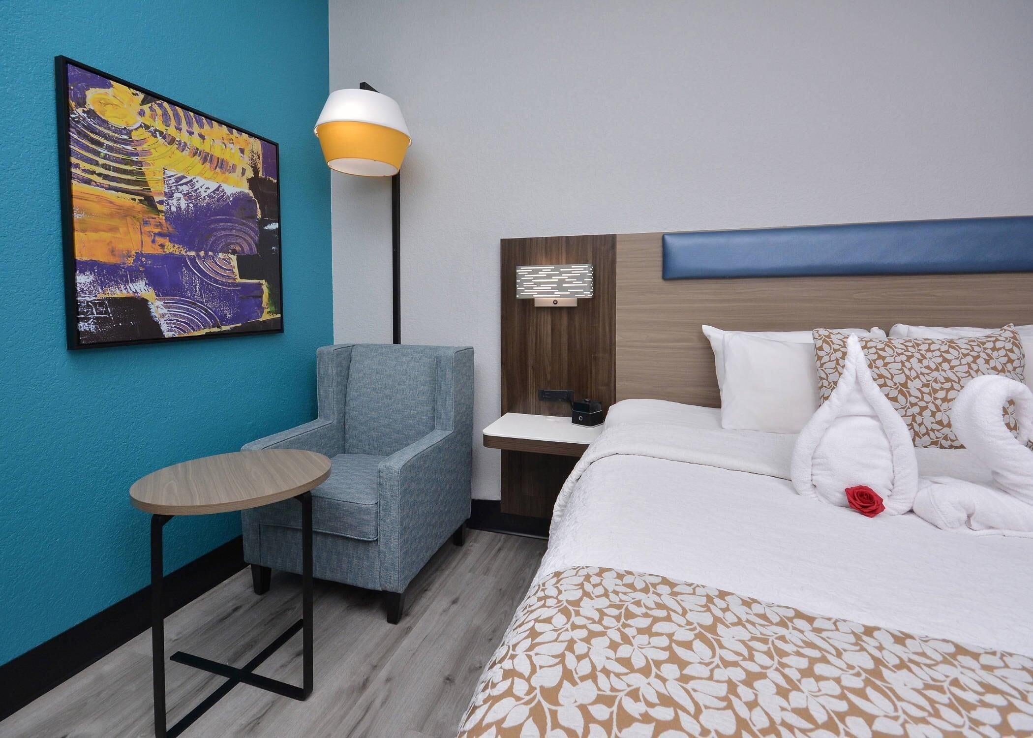 A hotel room with a bed, a chair, a small table, a lamp, and a painting on the wall.