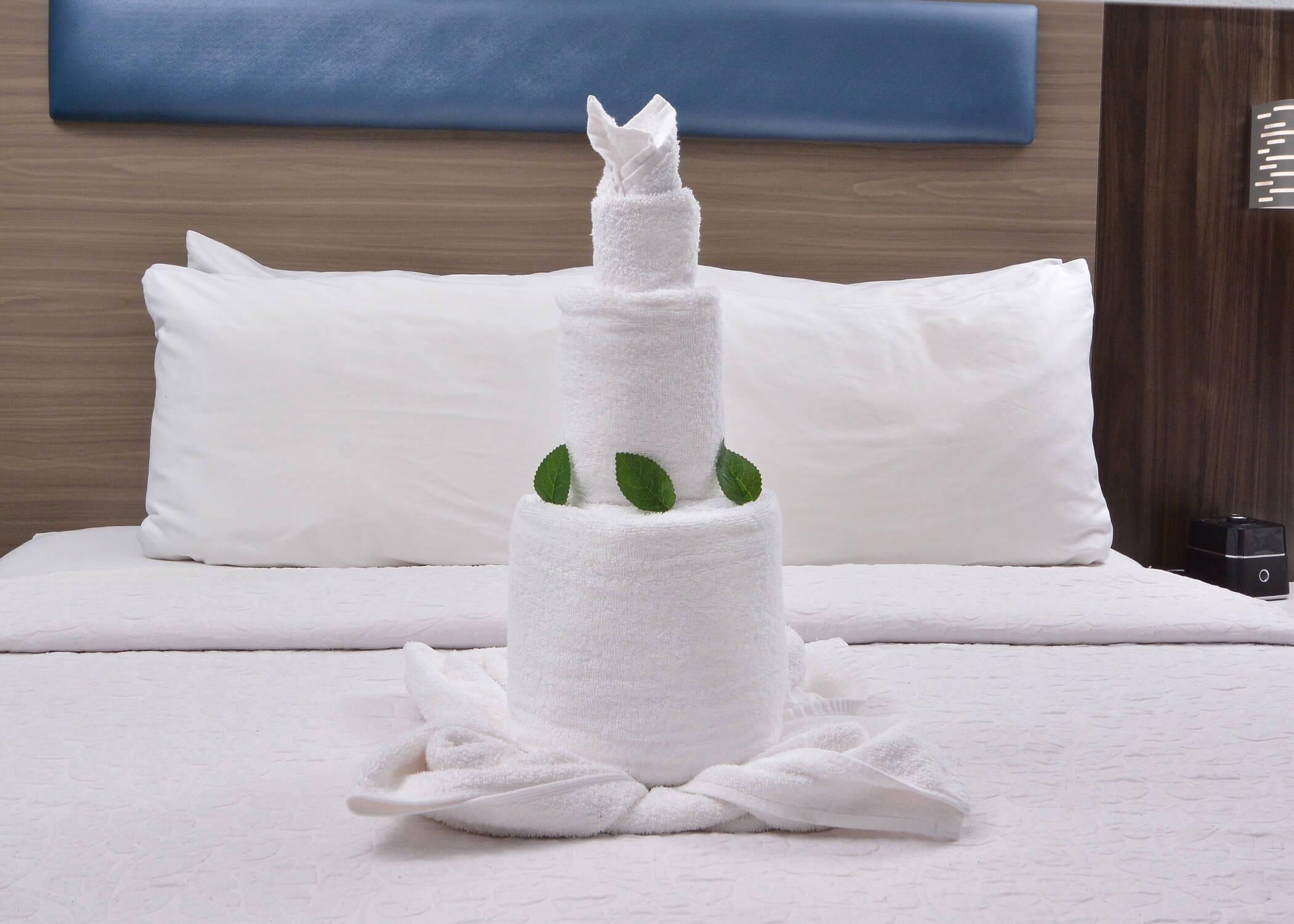 A bed with white bedding and pillows, a white towel sculpture, and a small black object on the side.