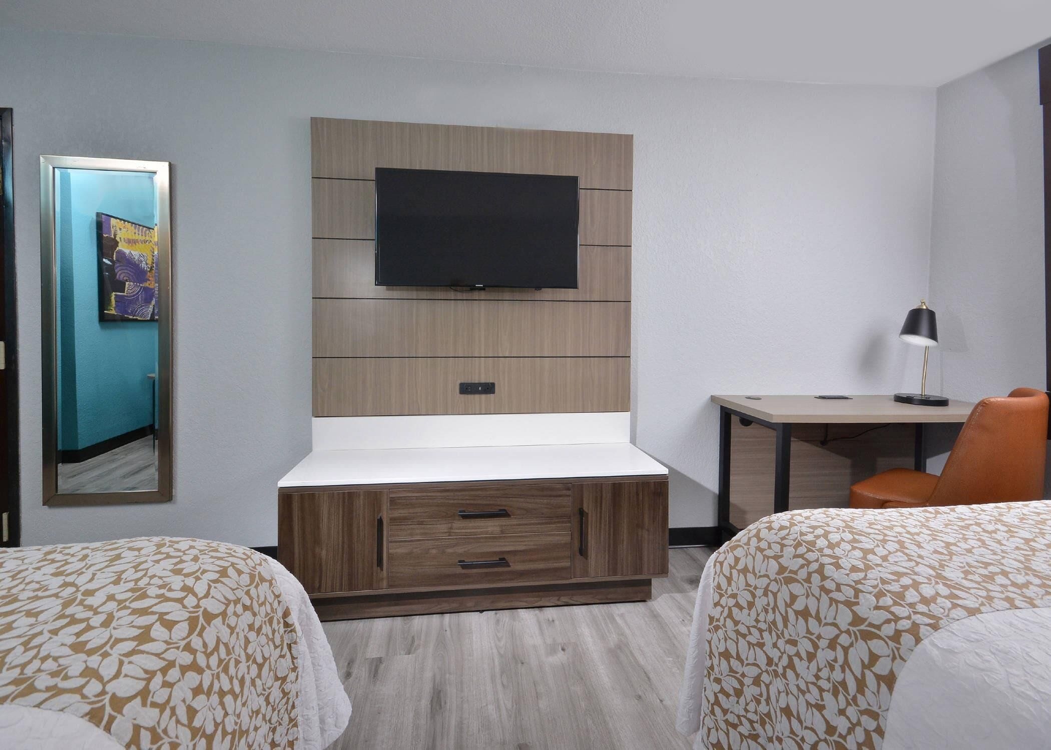 Habitación de hotel con dos camas, un televisor instalado en un panel de pared de madera, un escritorio con una silla y una lámpara.