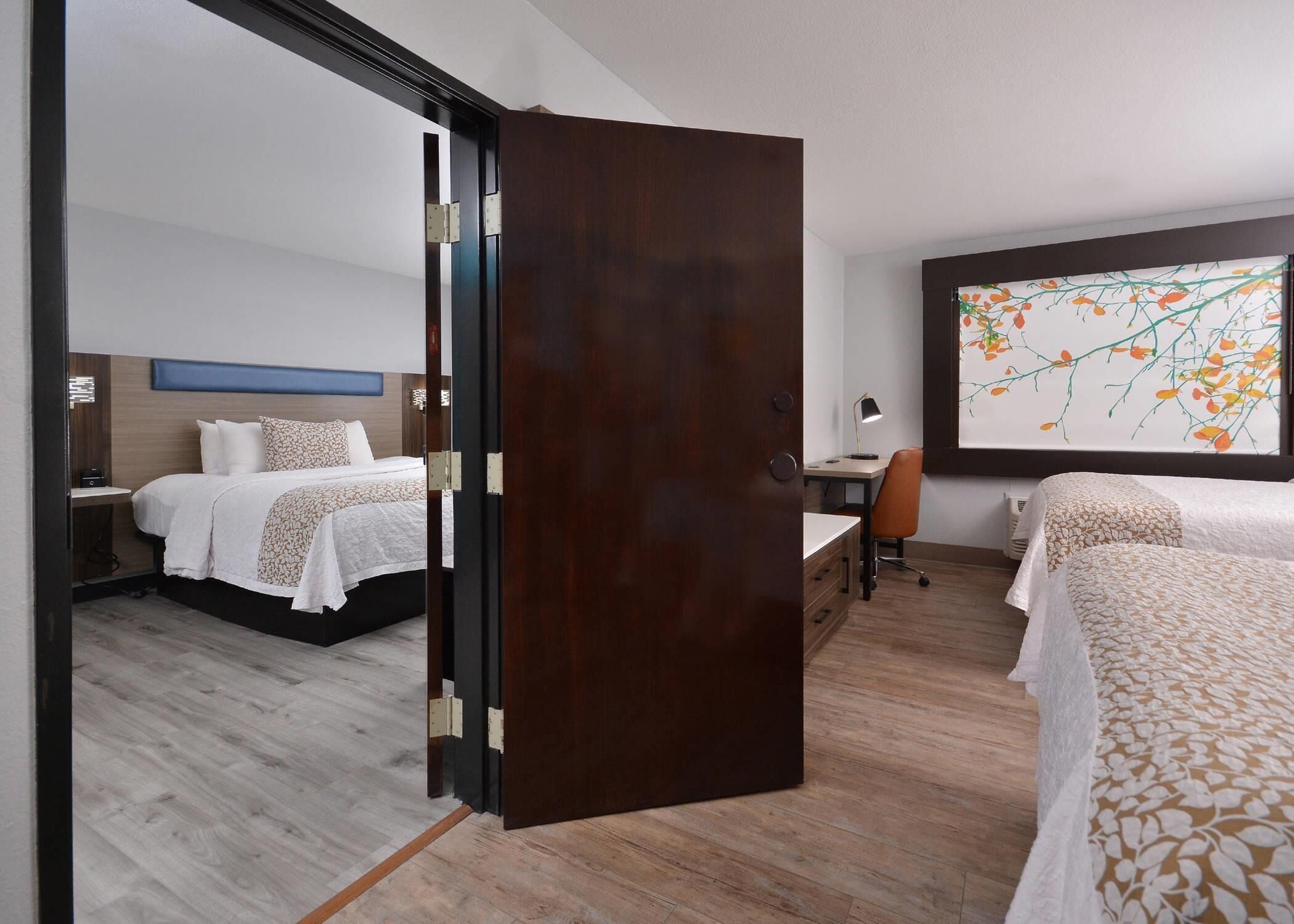 A hotel room with a bed, desk, chair, and a large window with a painting.