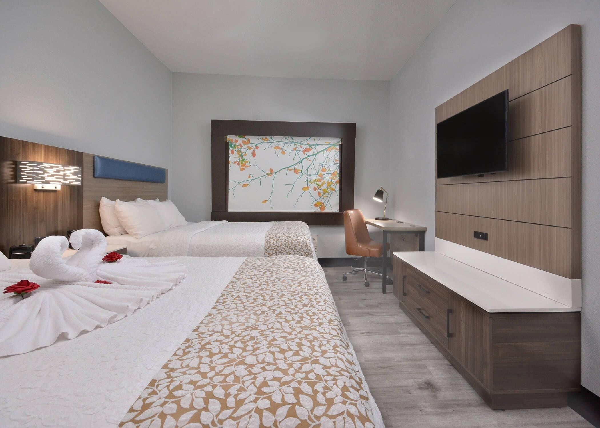 Habitación de hotel moderna con dos camas, un escritorio, una silla, un televisor y un amplio ventanal con una obra de arte floral.