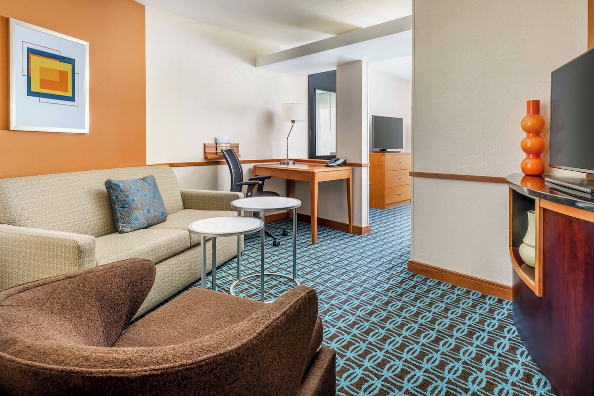 Habitación de hotel con sofá, sillón, pequeñas mesas redondas, un escritorio con silla, televisor y un jarrón decorativo.