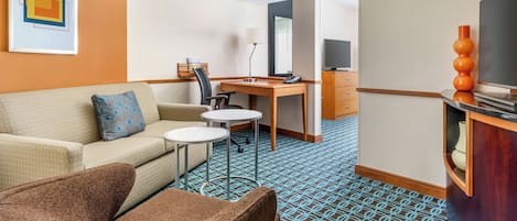 A hotel room with a sofa, armchair, small round tables, a desk with a chair, a TV, and a decorative vase.