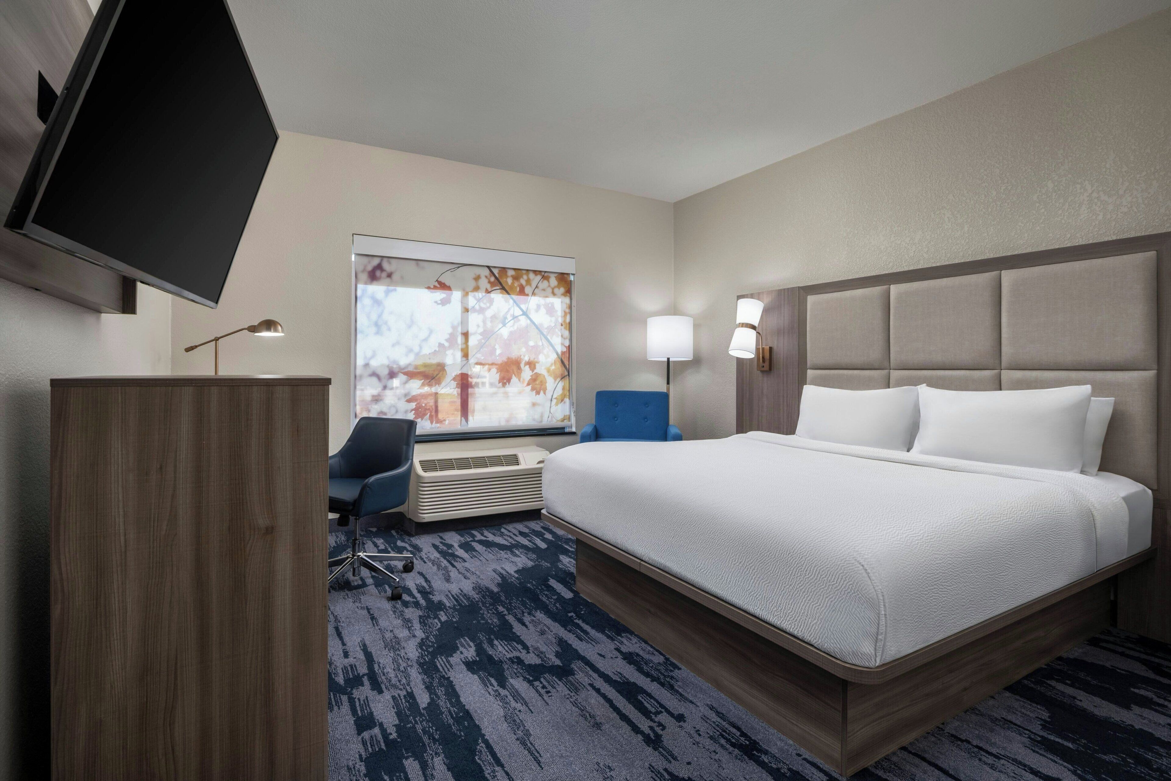 A hotel room with a large bed, a desk, a chair, a flat-screen TV, a window with a view, and a wall-mounted lamp.