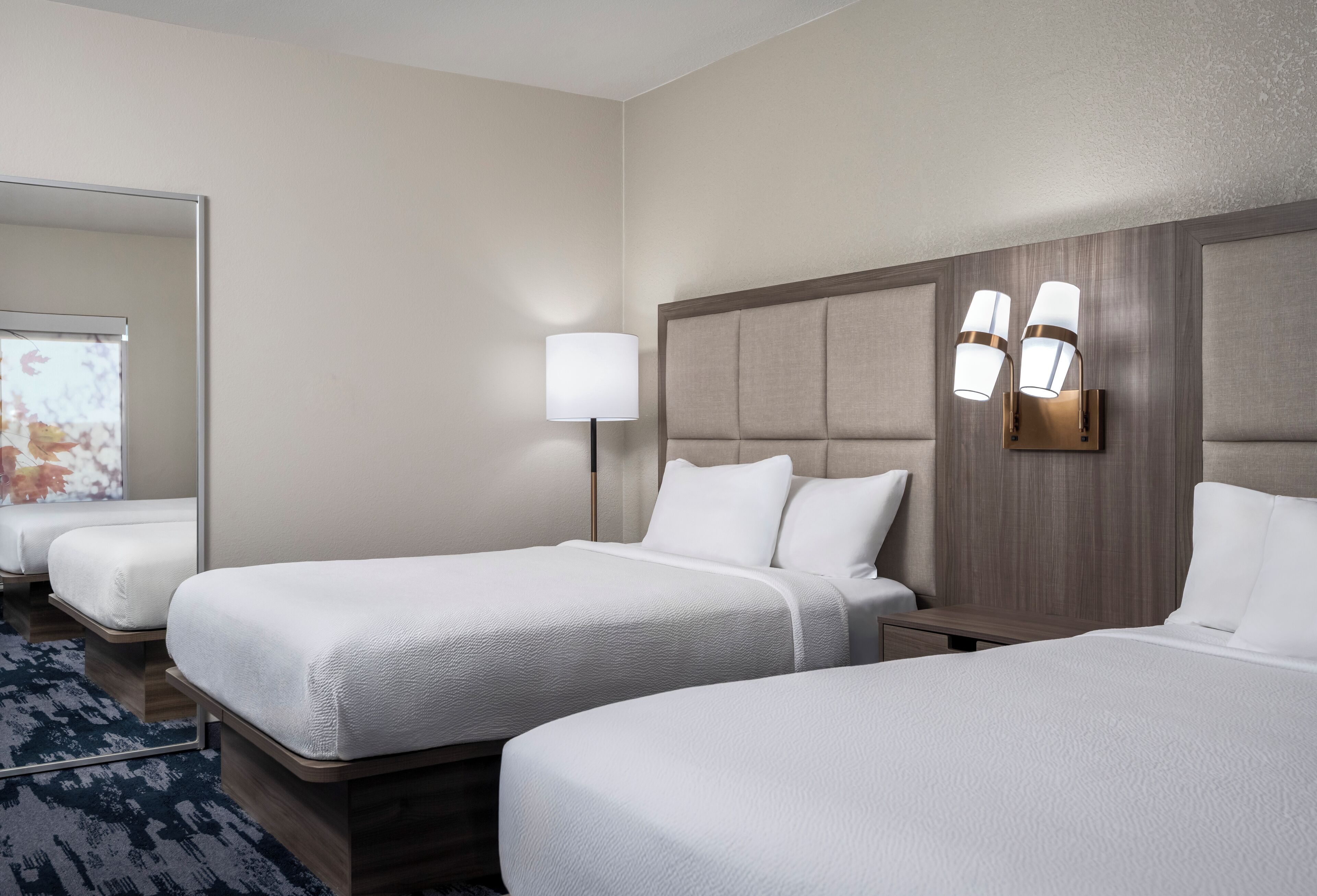 A hotel room with two beds, a large mirror, a wall-mounted lamp, and a carpeted floor.