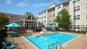 A swimming pool with lounge chairs and umbrellas, surrounded by a fence and a building with multiple windows. - Residence Inn by Marriott New Orleans Covington/North Shore (Covington)