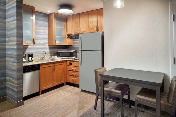 A compact kitchen with wooden cabinets, a stainless steel refrigerator, a microwave, and a coffee maker. There is a small dining table with two chairs.
