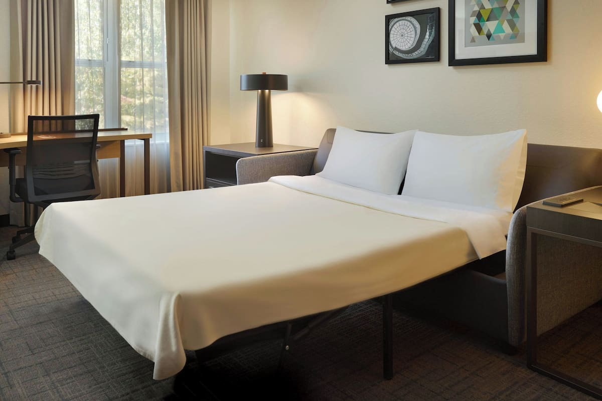 A hotel room with a neatly made bed, a desk with a chair, a lamp, and framed pictures on the wall.