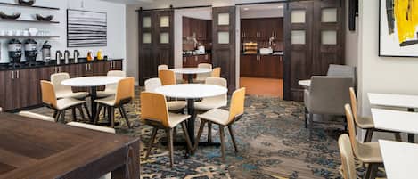 A hotel lobby with a central round table surrounded by chairs, a coffee station, and a view into the kitchen area.