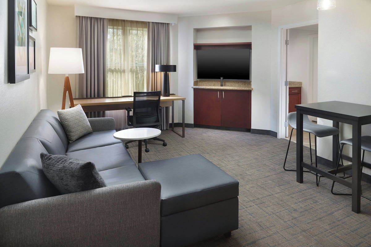 A modern hotel room with a grey sofa, a small round table, a desk with a chair, a flat-screen TV, and a kitchenette area.