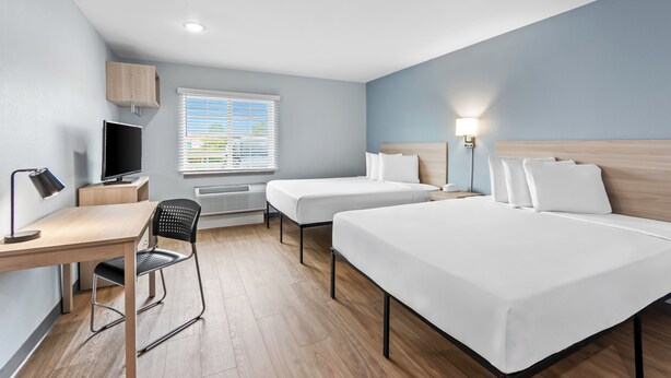A hotel room with a desk, two beds, a TV, and a window with blinds.