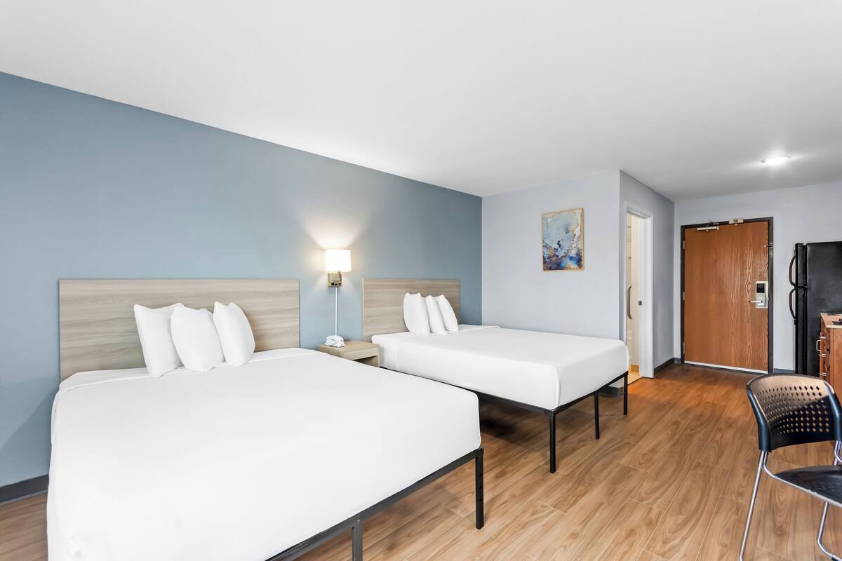 Hotellihuone, jossa on kaksi sänkyä, työpöytä, tuoli, jääkaappi ja taulu seinällä.