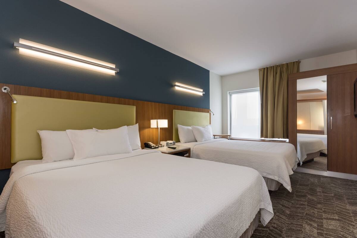 A hotel room with two beds, a wooden headboard, a nightstand with a lamp, and a large window with curtains.
