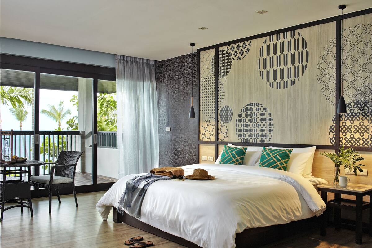 A modern bedroom with a large bed, a balcony with a table and chairs, and a view of palm trees.