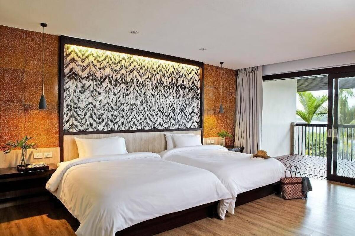 A hotel room with a large bed, a patterned headboard, a balcony with a view, and a nightstand with a lamp.