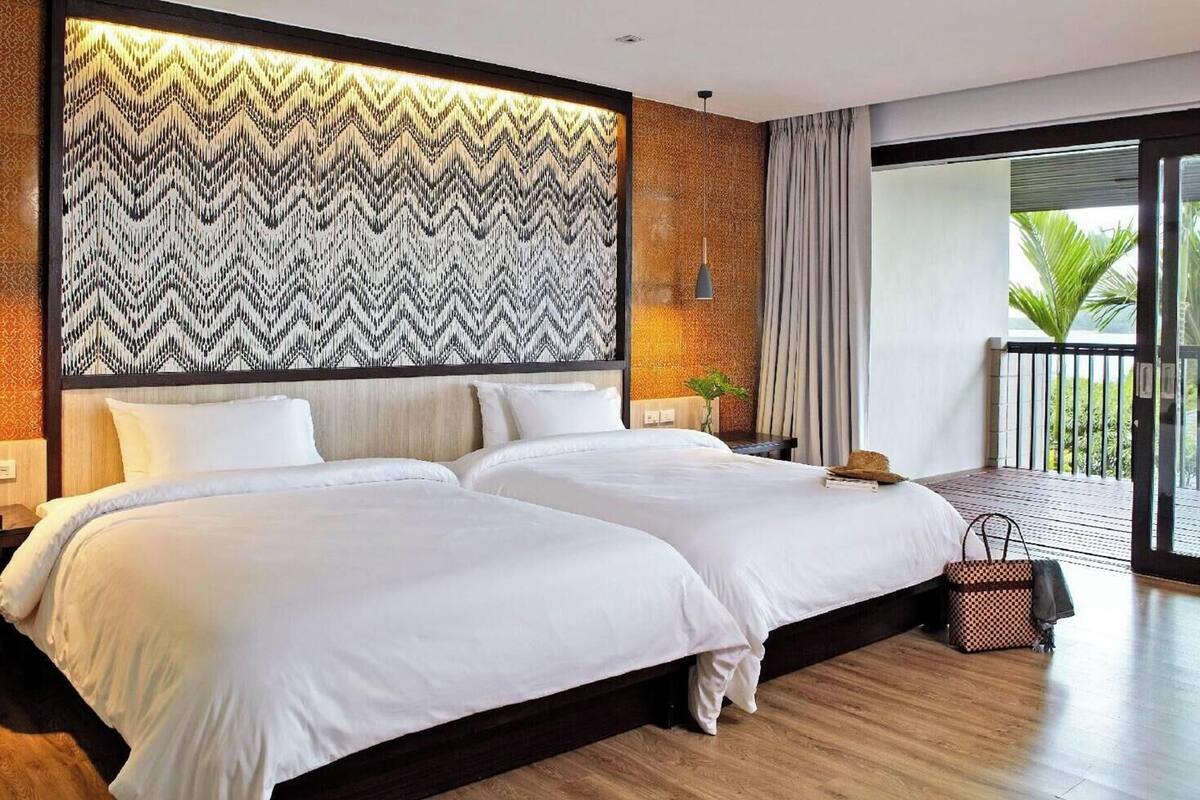 A double bed with a zigzag patterned headboard, a nightstand with a vase, and a view of a balcony with a palm tree.