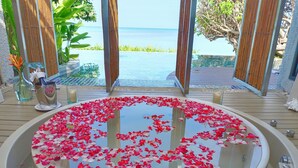 Una bañera de hidromasaje circular llena de agua y pétalos de rosa, rodeada por una terraza de madera y paneles de bambú, con vista al océano.