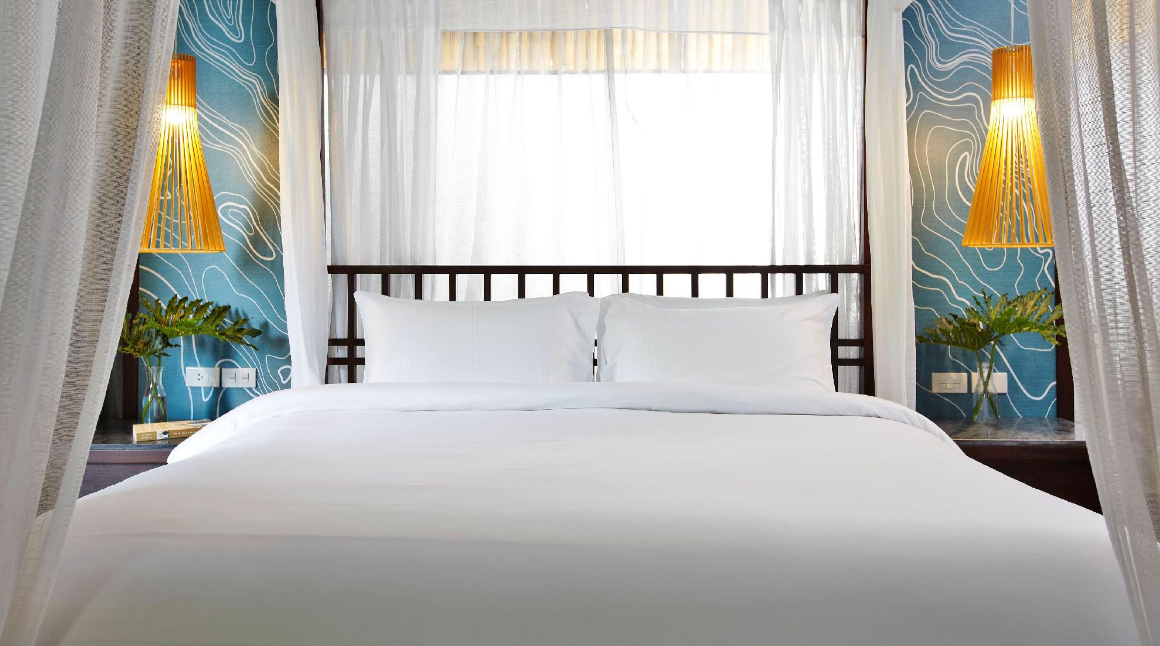 Una cama bien hecha con sábanas blancas en una habitación con paredes de diseño azul y cortinas blancas, y dos lámparas fijadas a la pared.