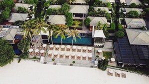 Vista aérea de un resort con piscina rodeada de sillas de descanso y sombrillas, palmeras y una playa.