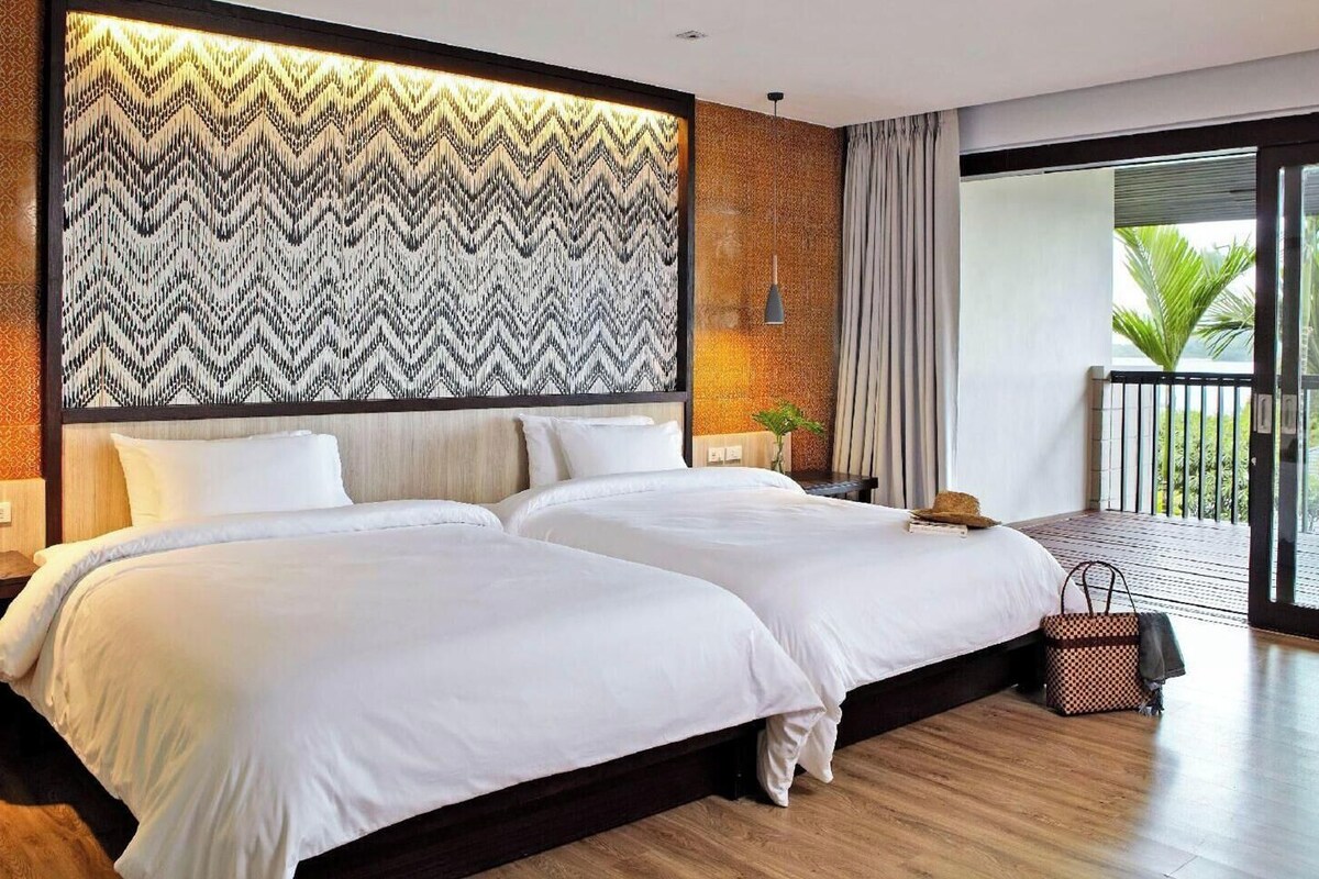Una cama doble con cabecera de estampado en zigzag, una mesita de noche con un florero, y vistas a un balcón con una palmera.