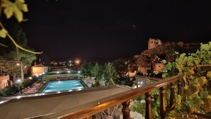 A nighttime view of a pool area with a wooden deck and lounge chairs, surrounded by greenery and a rocky cliff. - Elif Stone House (Ürgüp)