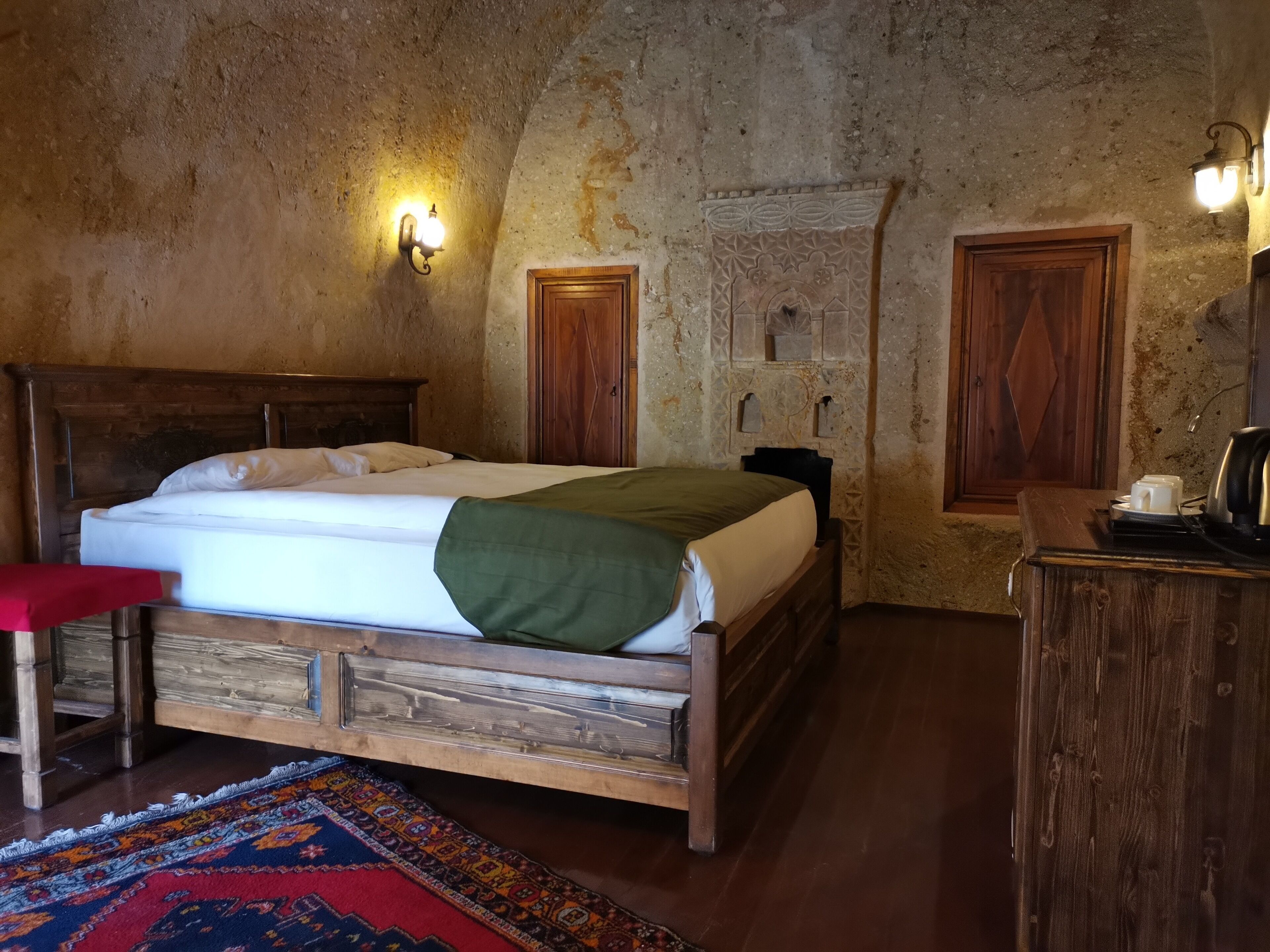 A bedroom with a wooden bed, a red bench, a patterned rug, and a stone wall with decorative carvings.
