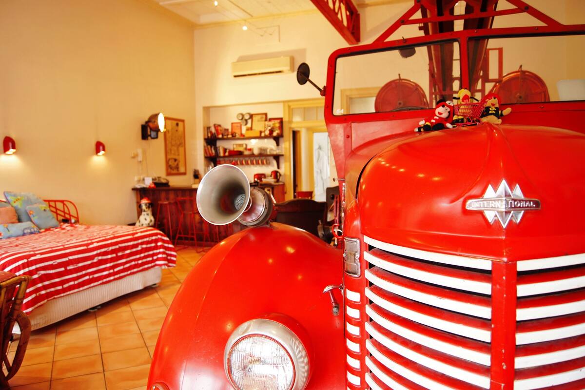 A room with a vintage red fire truck as a centerpiece, a bed with a striped blanket, and a small table with a chair.