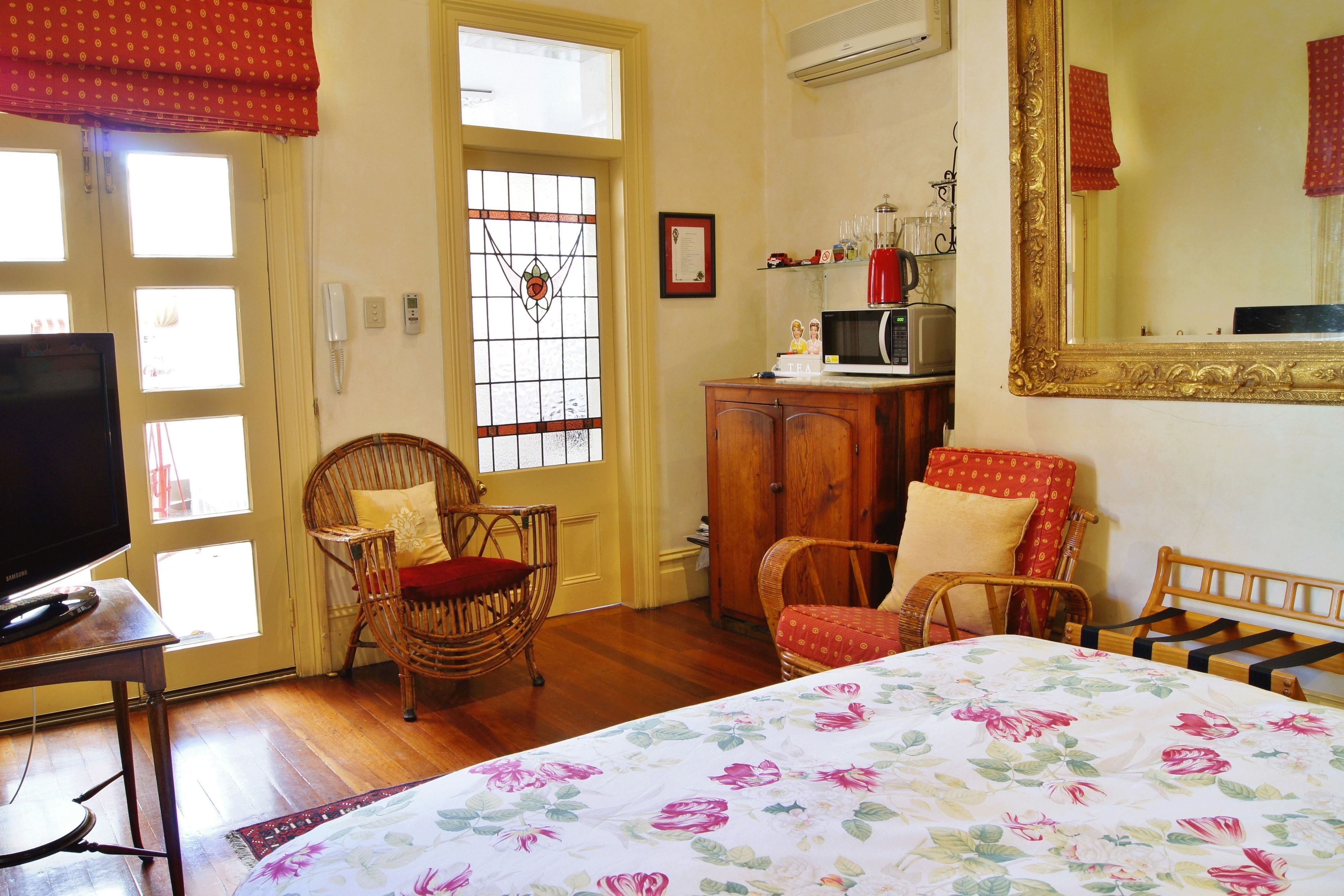 Una habitación con una cama, dos sillones de mimbre, un televisor, un armario, un microondas y un espejo grande.