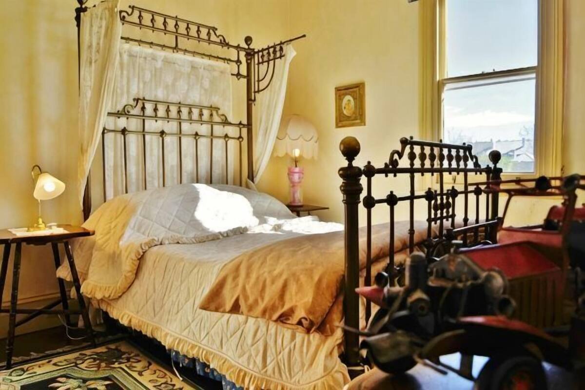 A bedroom with a four-poster bed, a bedside table with a lamp, a nightstand, and a window with a view.