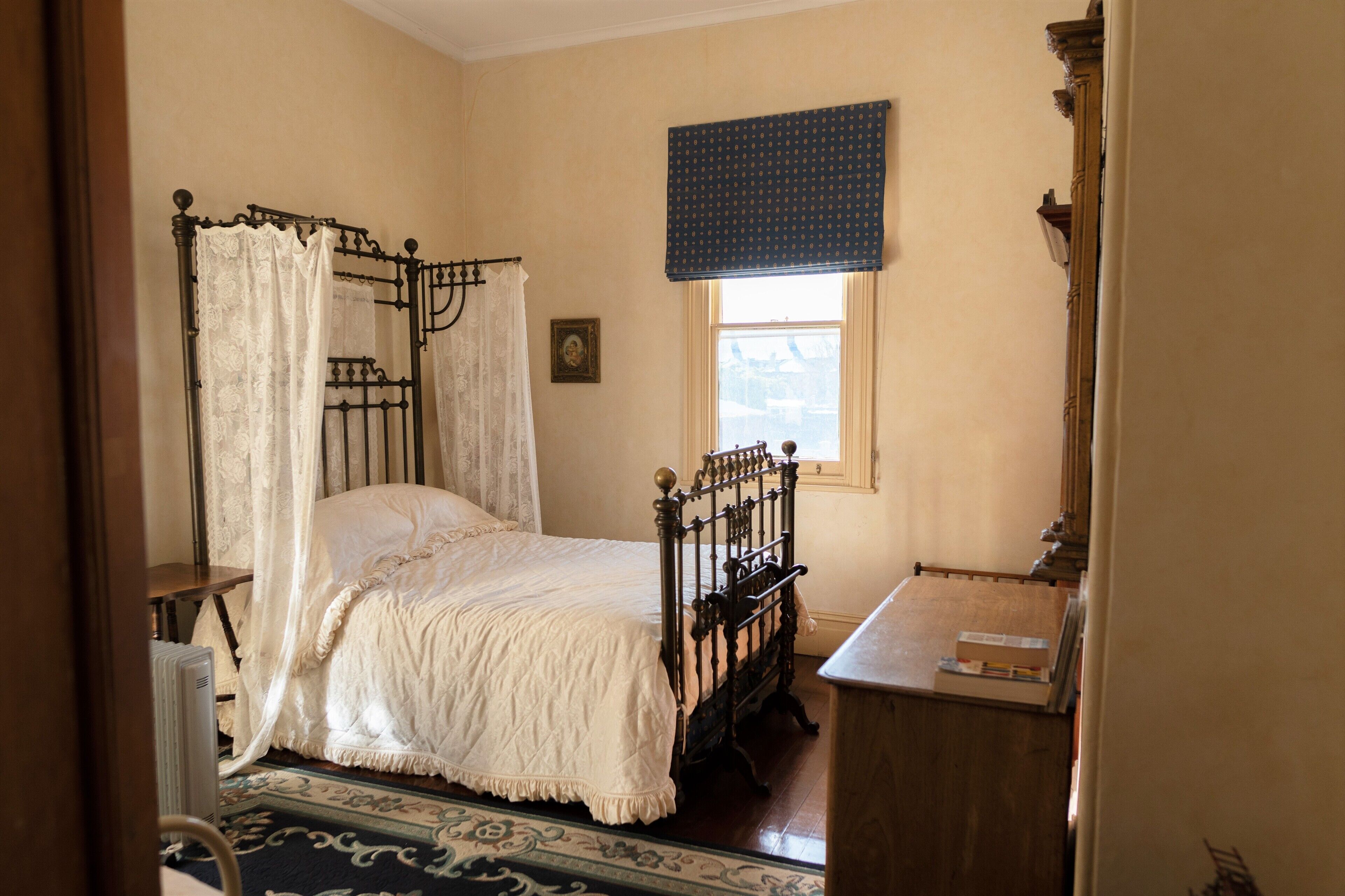 Un dormitorio con una cama con dosel, una mesita de noche de madera y una ventana con un visillo azul.
