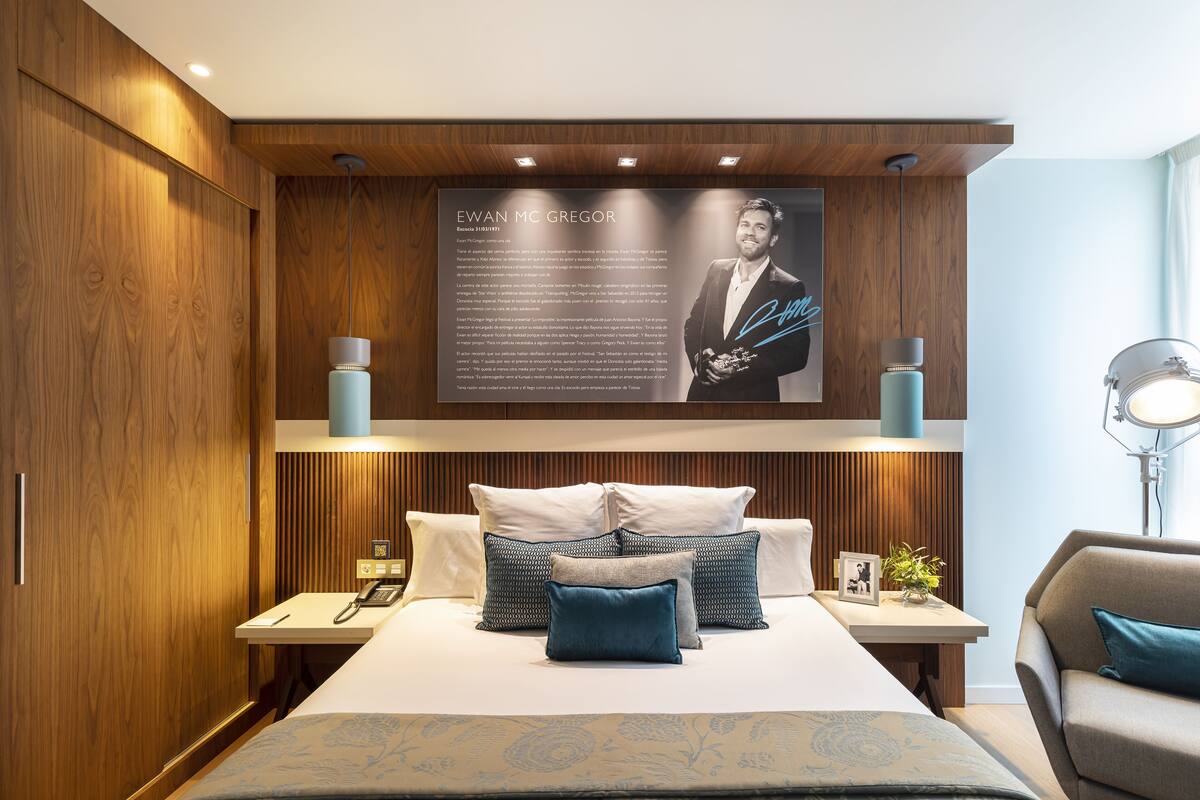 A modern hotel room with a large bed, wooden paneling, and a framed portrait above the bed.