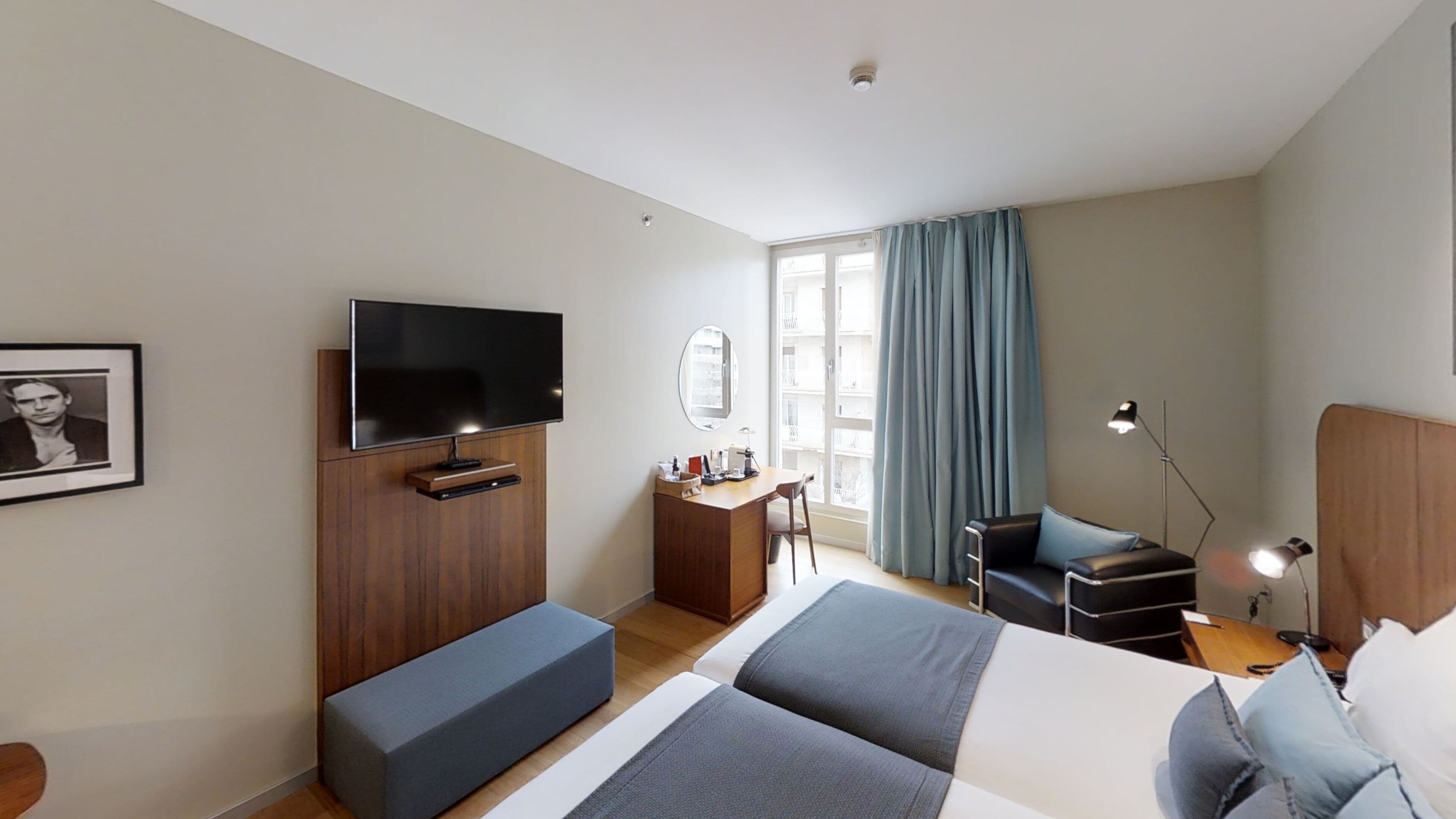 Une chambre d’hôtel comprenant un lit, un bureau, une chaise, un téléviseur, une fenêtre avec des rideaux et un tableau encadré au mur.