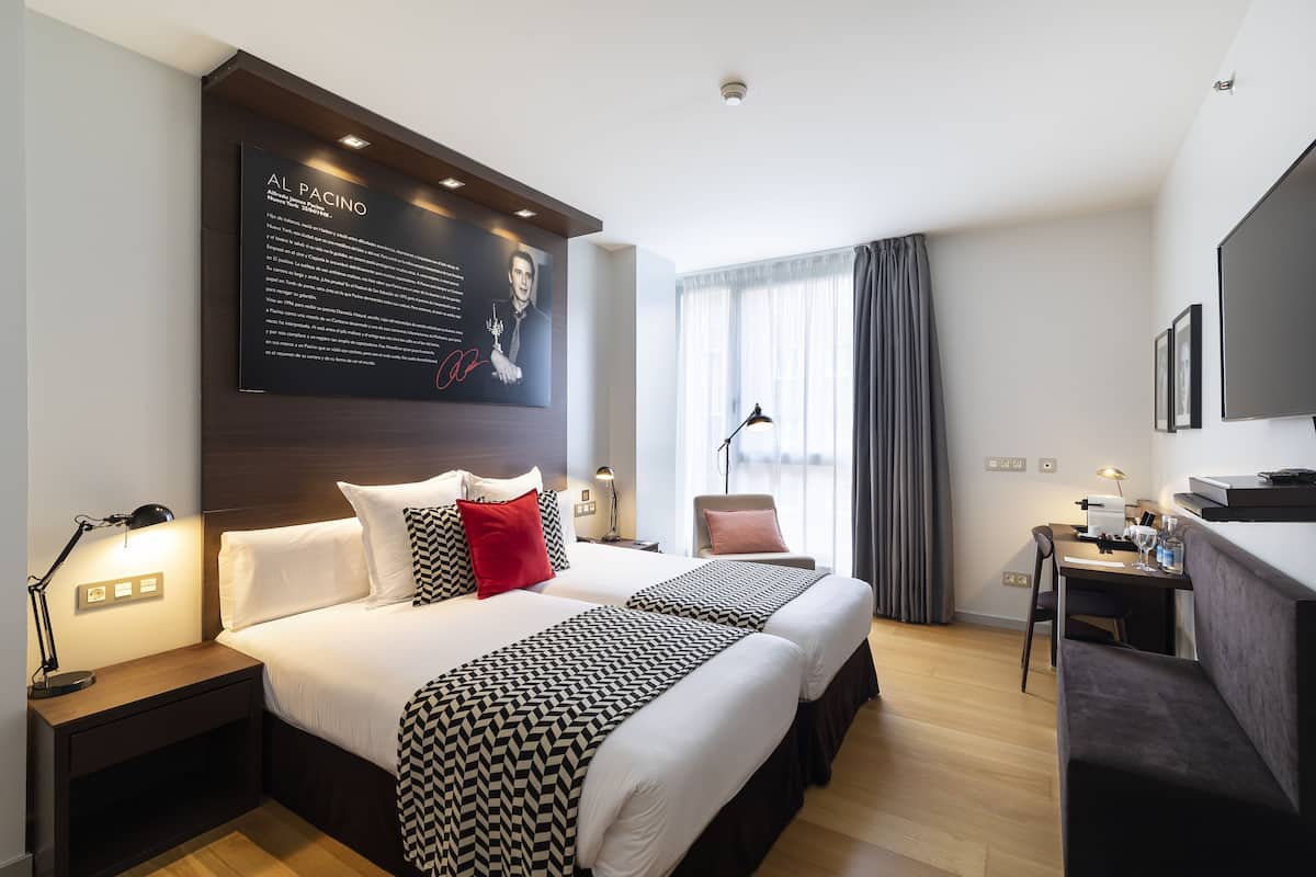 A modern hotel room with two beds, a desk, a chair, and a large portrait on the wall.