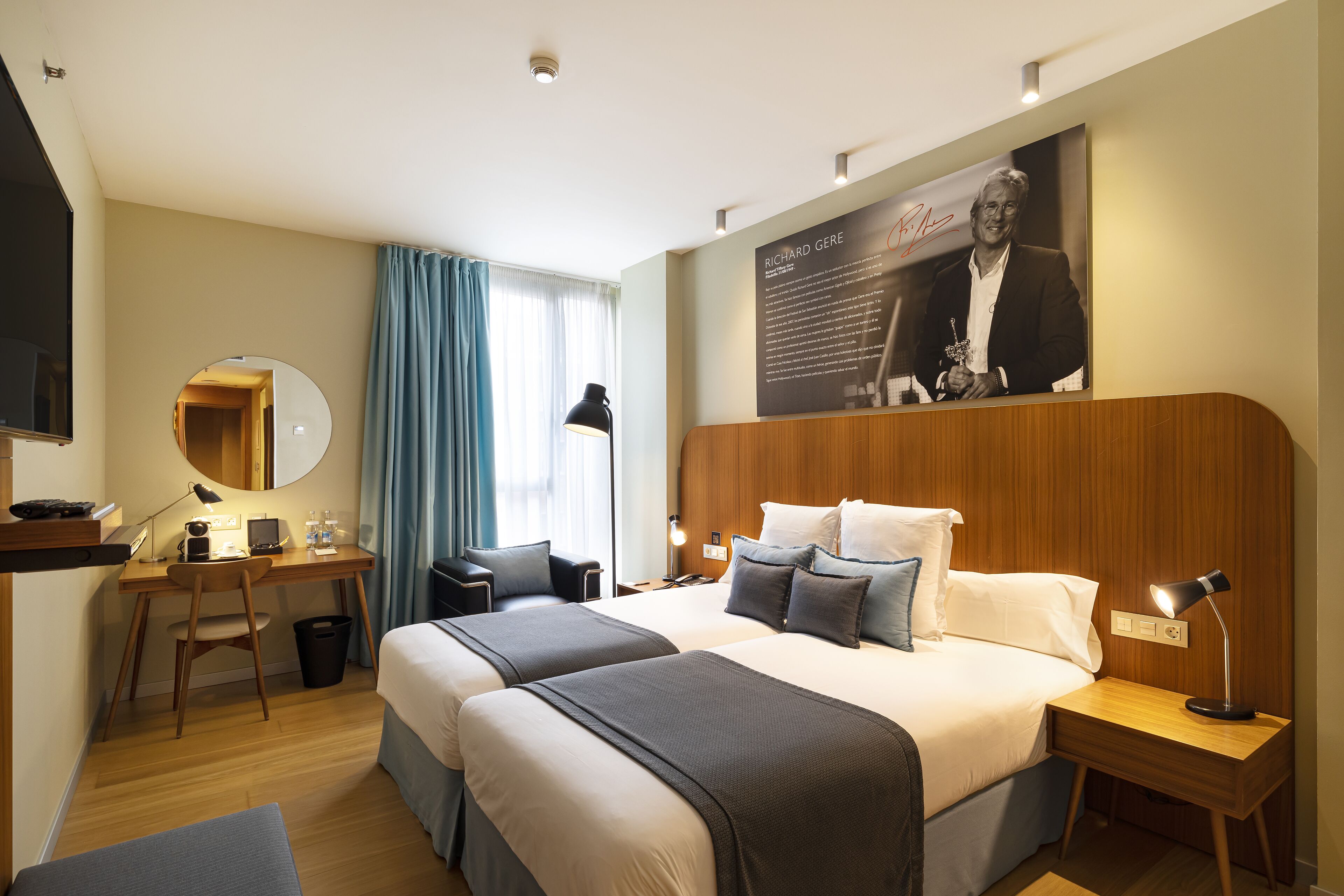 Camera d'albergo con due letti, una scrivania, una sedia, una televisione, un quadro appeso al muro e un comodino con una lampada.
