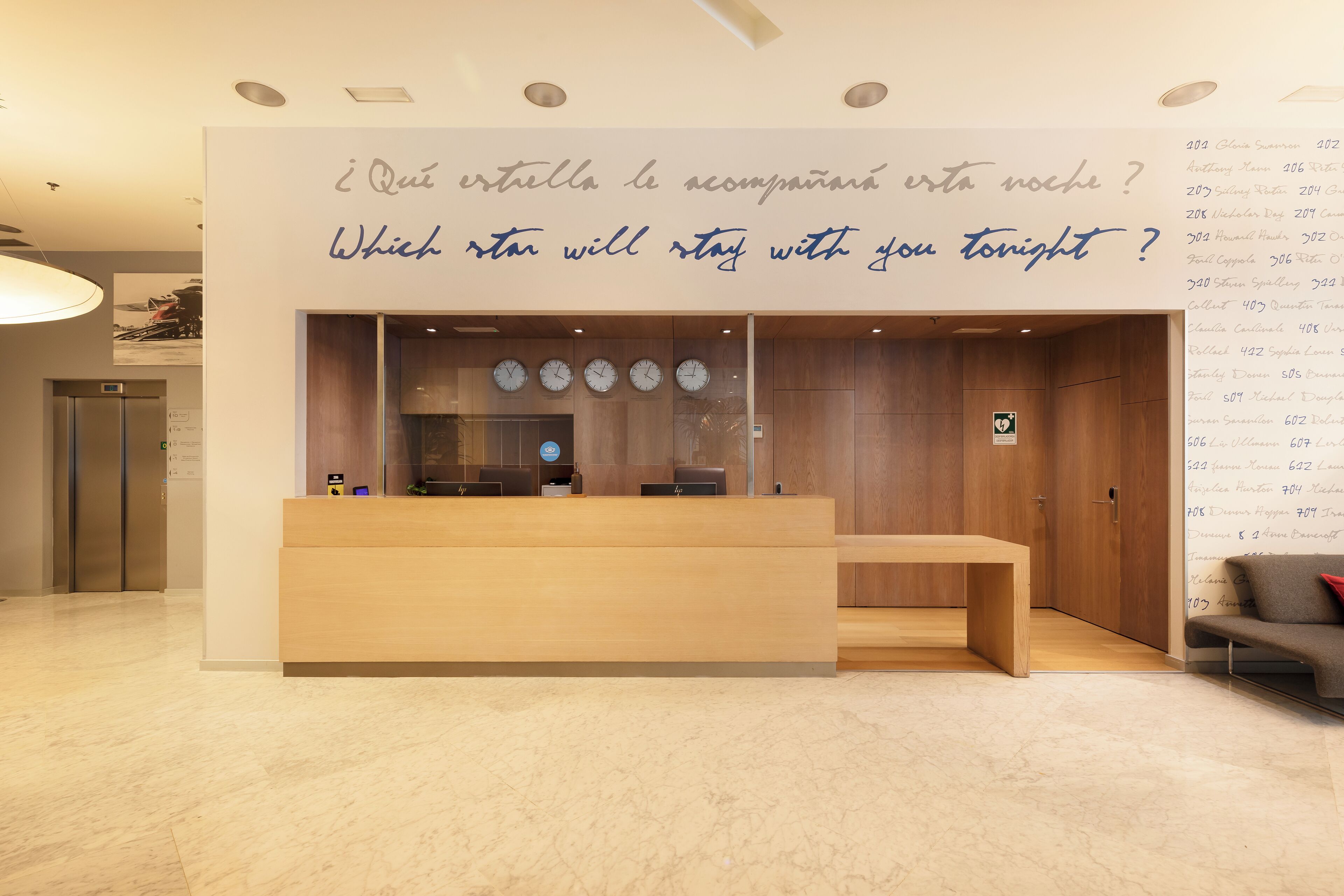 A hotel reception area with a wooden reception desk, a wall with a question in two languages, and a list of room numbers on the right.
