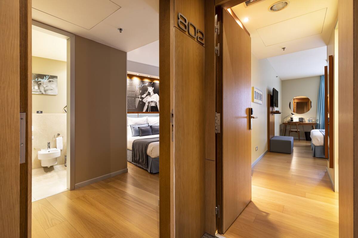 A hotel room with a wooden floor, a bed with a gray headboard, a bedside table with a lamp, a small desk with a chair, and a bathroom with a sink and toilet.
